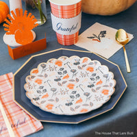 Close-up place setting with layered plates, plaid “Grateful” cup, white-pumpkin napkin, and gold spoon.