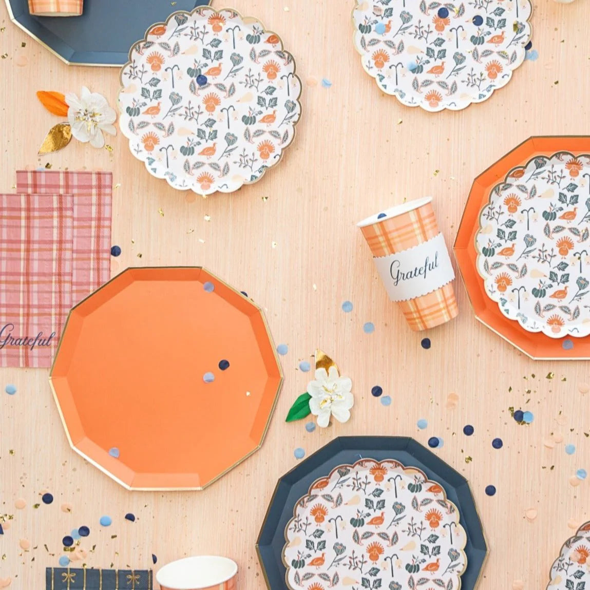 Alt: Flat lay of octagon plates, floral salad plates, plaid “Grateful” cup, and guest towels with confetti on peach background.