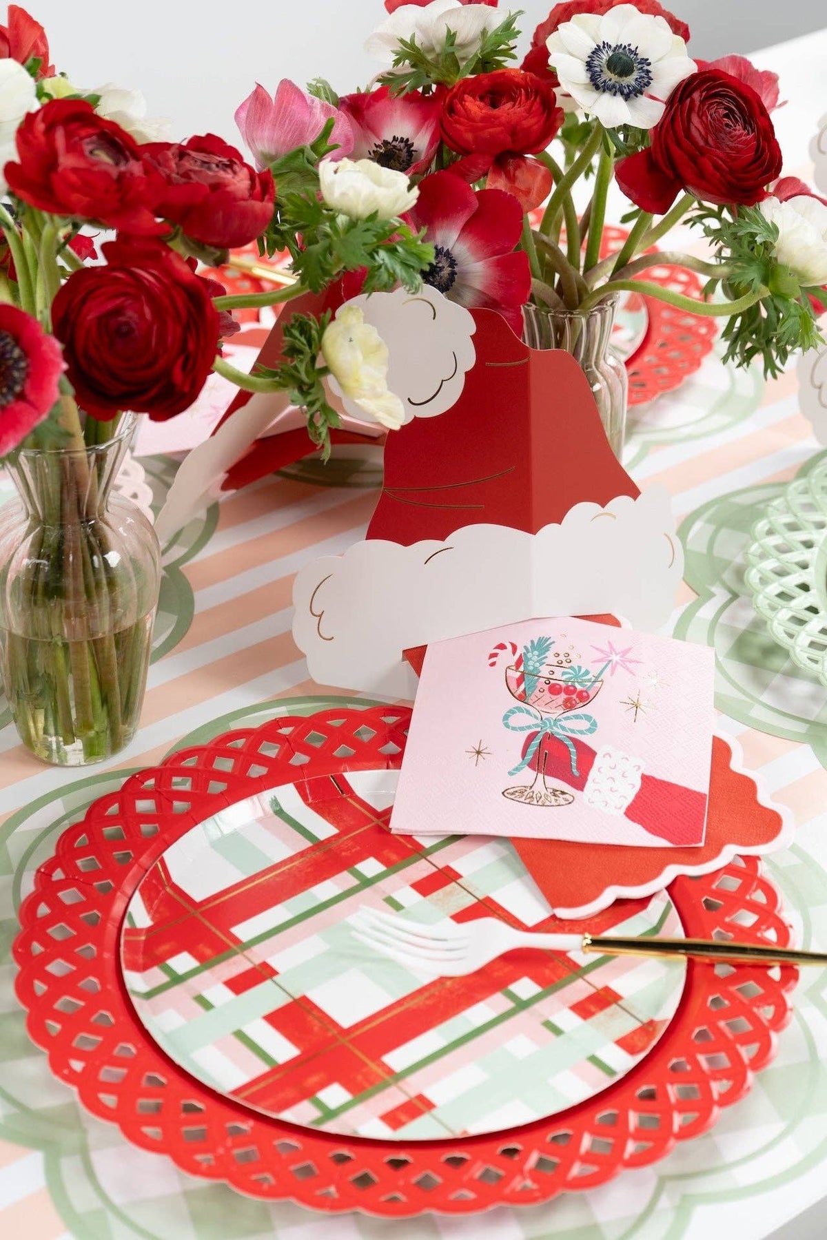 Place setting with a green lattice charger, red plaid plate, a folded red Santa-hat card, a pink present-print guest towel, and flowers in vases.