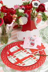Place setting with a green lattice charger, red plaid plate, a folded red Santa-hat card, a pink present-print guest towel, and flowers in vases.