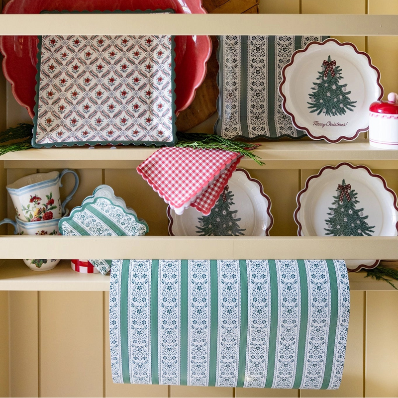 Green floral stripe paper table runner draped from a cream kitchen hutch below Christmas tree plates, red gingham napkins, and square floral plates