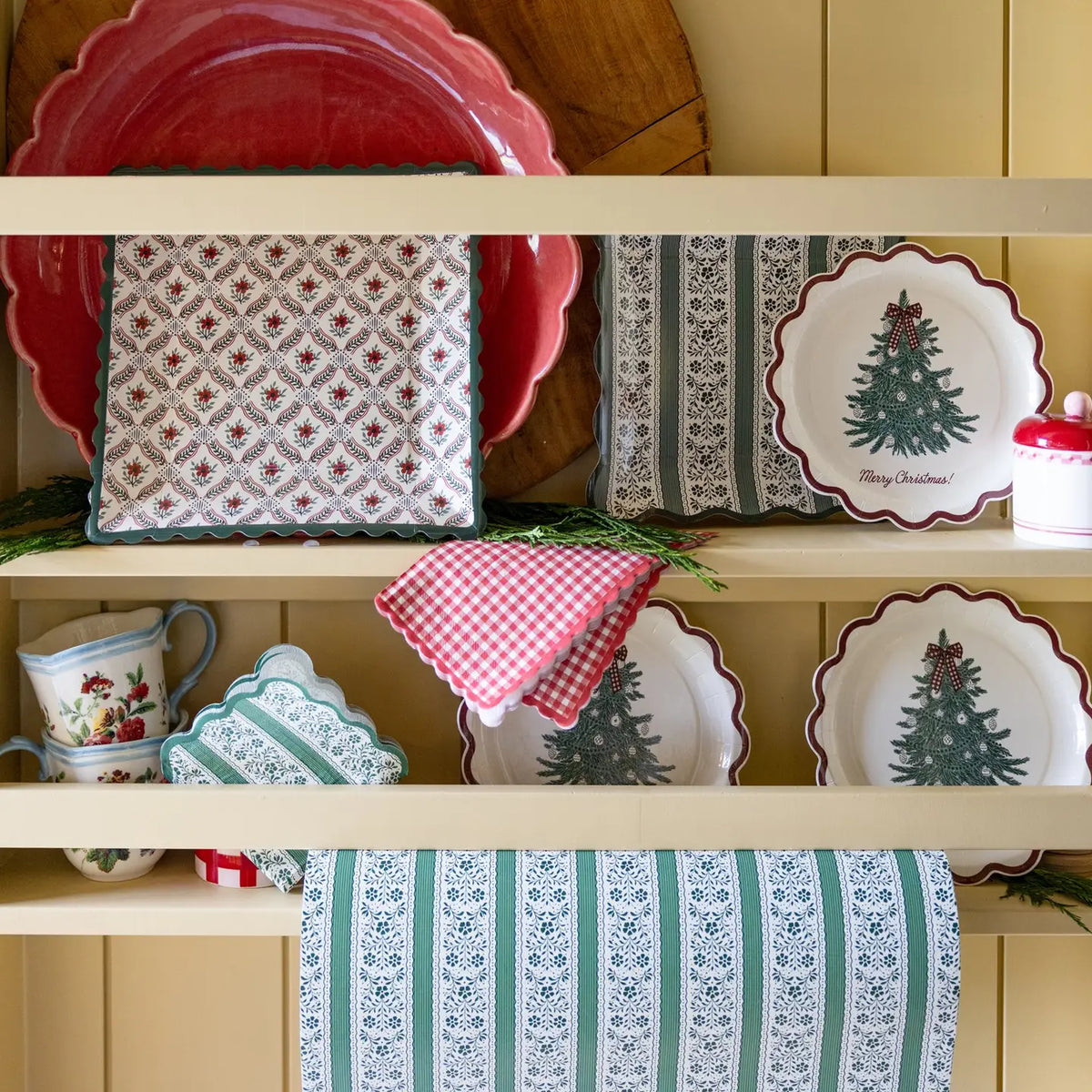 Green floral stripe square paper plates on a cream kitchen hutch with Christmas tree plates, red gingham napkins, and cedar greenery