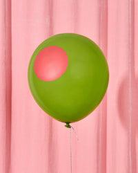 Single green olive latex balloon with red pimento circle floating in front of pink curtain backdrop