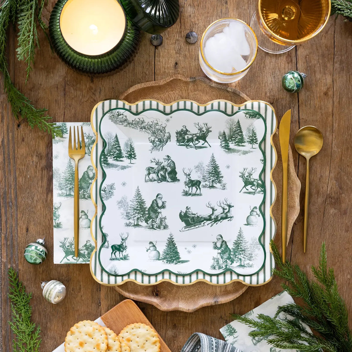Green ticking stripe square paper plate with a gold foil edge layered under a green toile Santa plate at a Christmas place setting with gold flatware and a candle