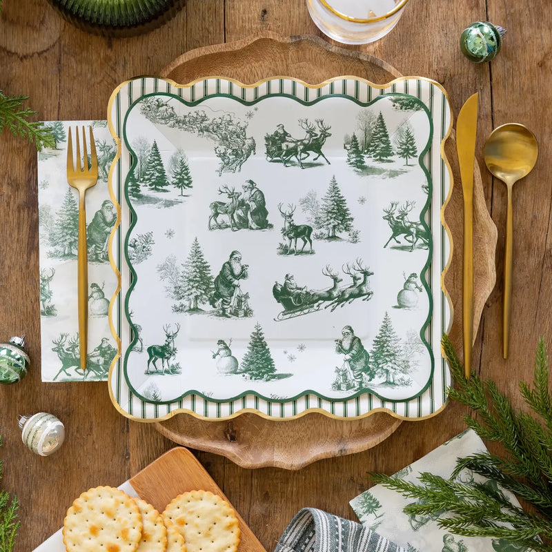 Green toile Santa paper dinner napkin under a gold fork beside a green toile scalloped square plate on a wooden table with ornaments and greenery