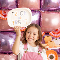 Smiling child in pink overalls holds “TRICK OR TREAT” plate aloft against metallic balloon wall, demonstrating fun photo-prop potential.