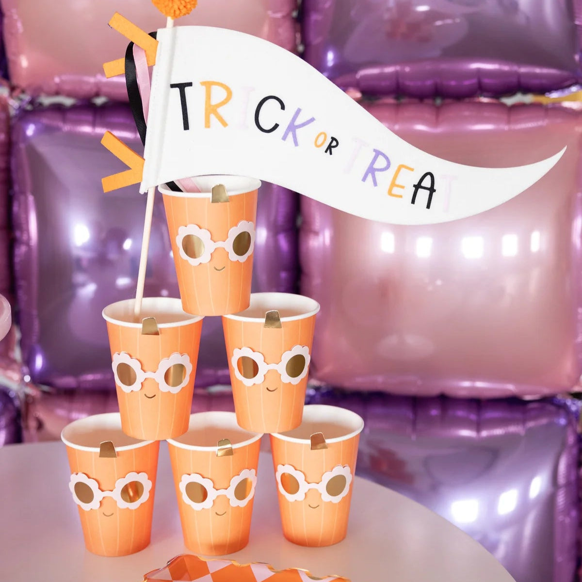 Close-up of six Groovy Pumpkin cups stacked beneath felt “Trick or Treat” flag, highlighting daisy-shape sunglasses and gold accents.