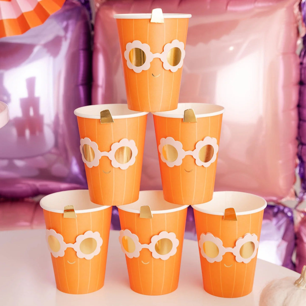 Pyramid of Groovy Pumpkin cups on party table; metallic sunglasses design and gold stems pop against pink-purple balloon wall and striped fans.