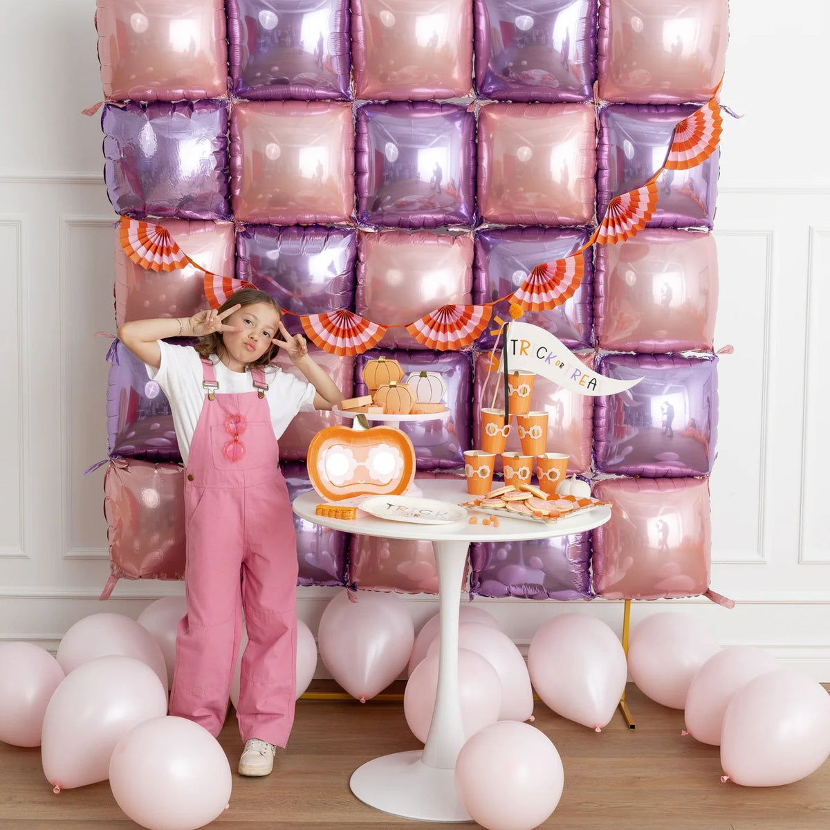 Wide scene of balloon square backdrop with girl posing beside table stacked with Groovy Pumpkin cups, treat boxes and Halloween décor.