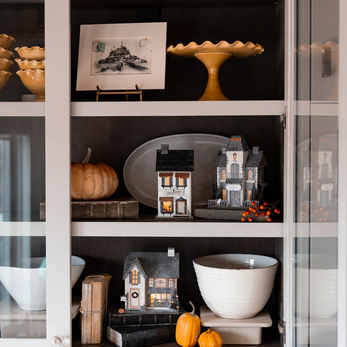 Styled bookshelf with lighted Halloween village houses including the Haberdashery shop glowing beside pumpkins and berries.
