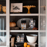 Styled bookshelf with lighted Halloween village houses including the Haberdashery shop glowing beside pumpkins and berries.