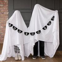 Lifestyle shot: two children hidden beneath flowing white bed sheets stand side by side like playful phantoms; black half-moon felt pennants with bold white letters and pearl-cream pom-poms swoop from one “ghost” to the other, repeating BOO across their fronts. Brick wall and charcoal wainscoting backdrop add texture, while striped stockings and checkered sneakers peek out, illustrating banner length, flexibility and family-friendly Halloween fun.