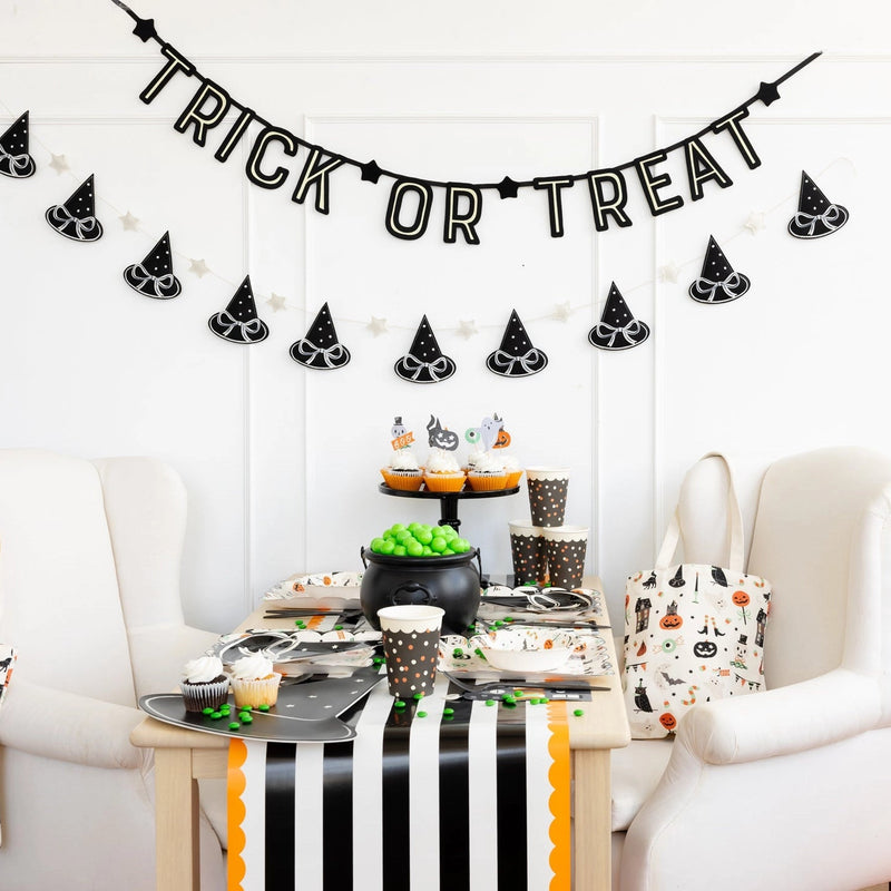Black felt ‘TRICK OR TREAT’ halloween banner hanging above a witchy cupcake table with striped runner