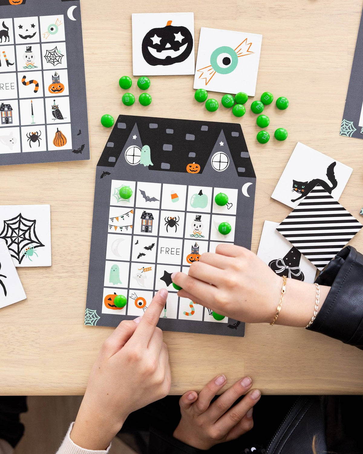 Kids playing Halloween bingo, marking a haunted house bingo sheet with candy while calling cards sit nearby
