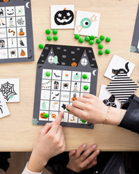 Kids playing Halloween bingo, marking a haunted house bingo sheet with candy while calling cards sit nearby