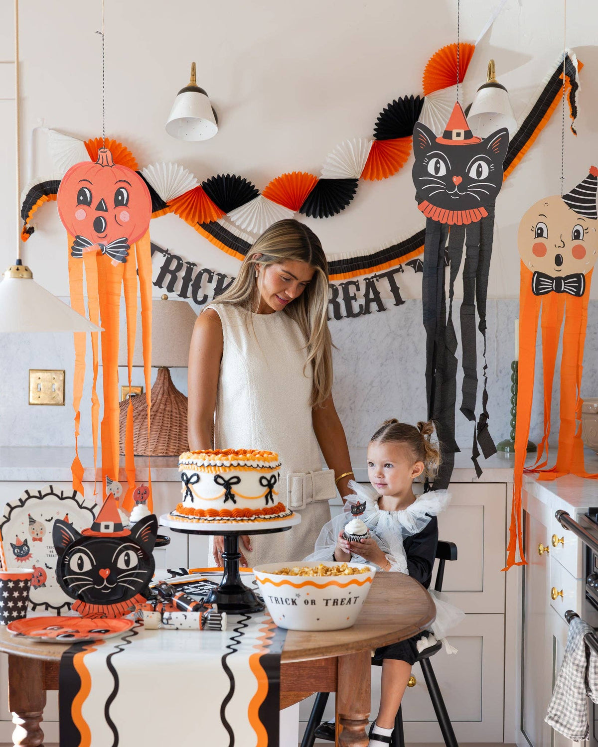 Woman and child in a kitchen decorated for Halloween with a cake and decorations.
