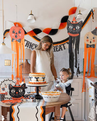 Woman and child in a kitchen decorated for Halloween with a cake and decorations.
