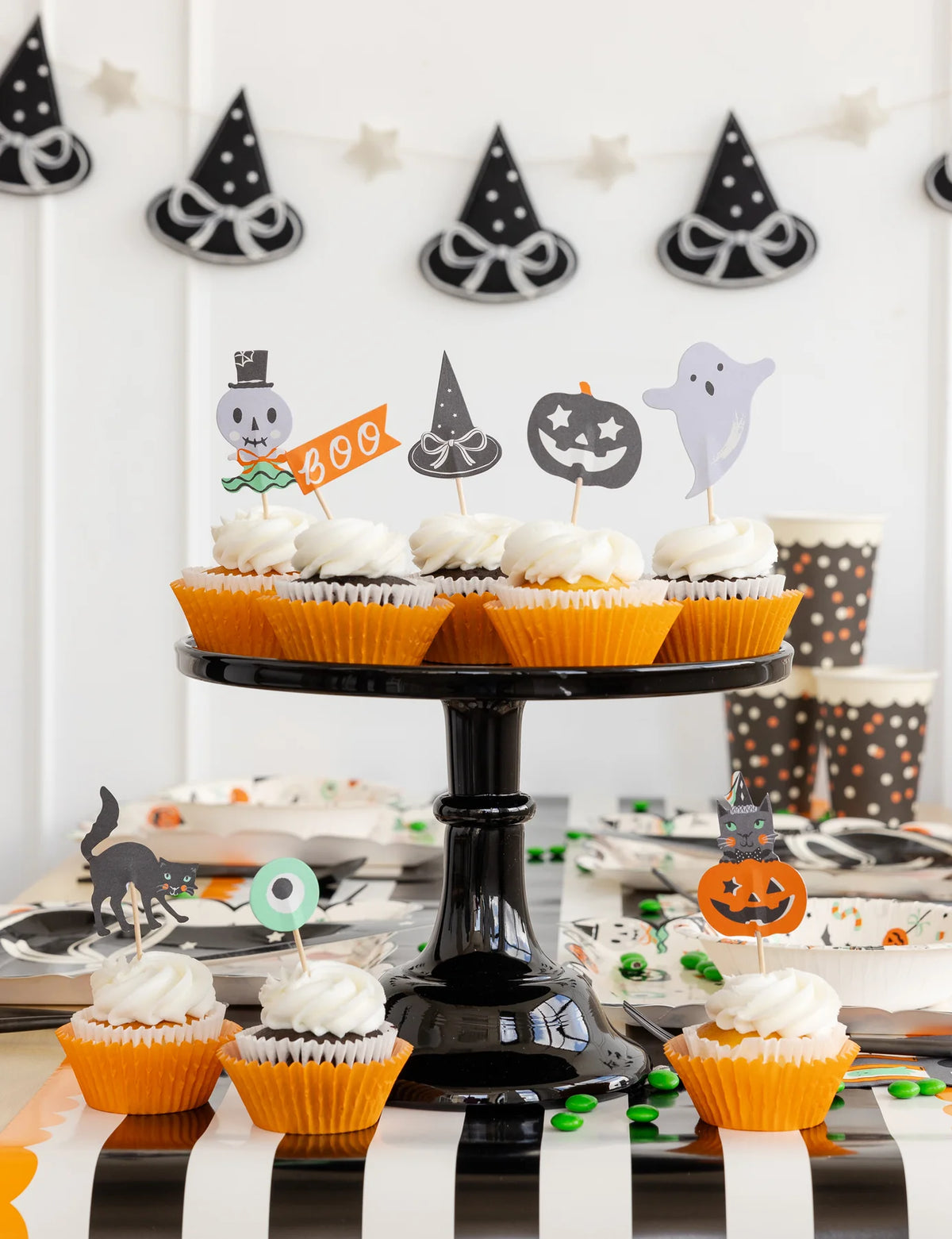 Close-up of cupcakes on black stand topped with Halloween picks; witch-hat banner blurred in background.