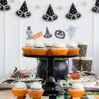 Halloween-themed cupcakes on a stand with decorative toppers against a white wall with black witch hats.
