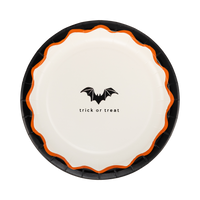 Cream Halloween dessert plate with a black bat icon and "trick or treat" text, black outer rim and orange scalloped inner edge, overhead view on gray background.