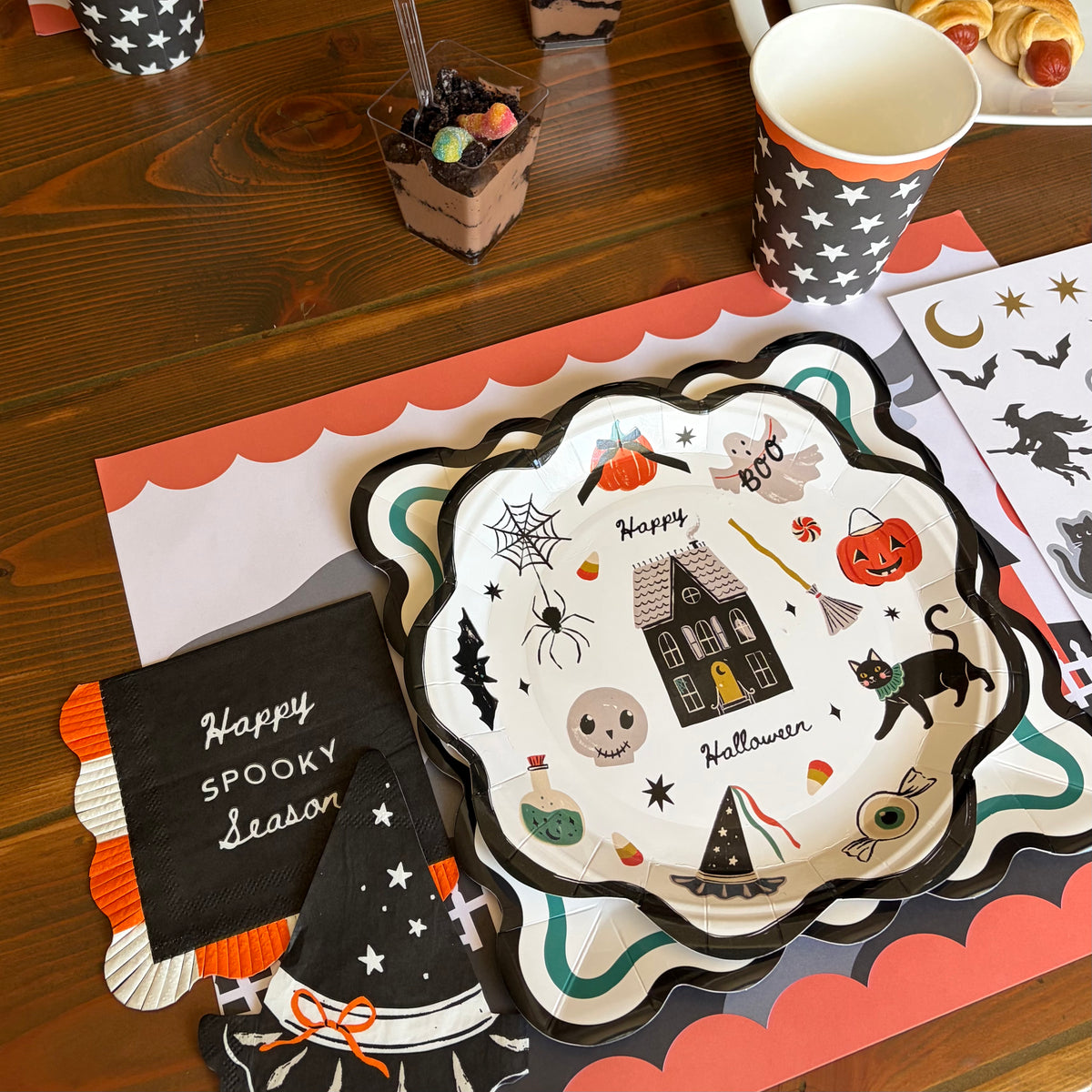 Halloween place setting with a scalloped icon placemat, a Happy Spooky Season chalkboard sign, an icon plate with ghosts, cats, and a haunted house, and a black star cup on a wood table