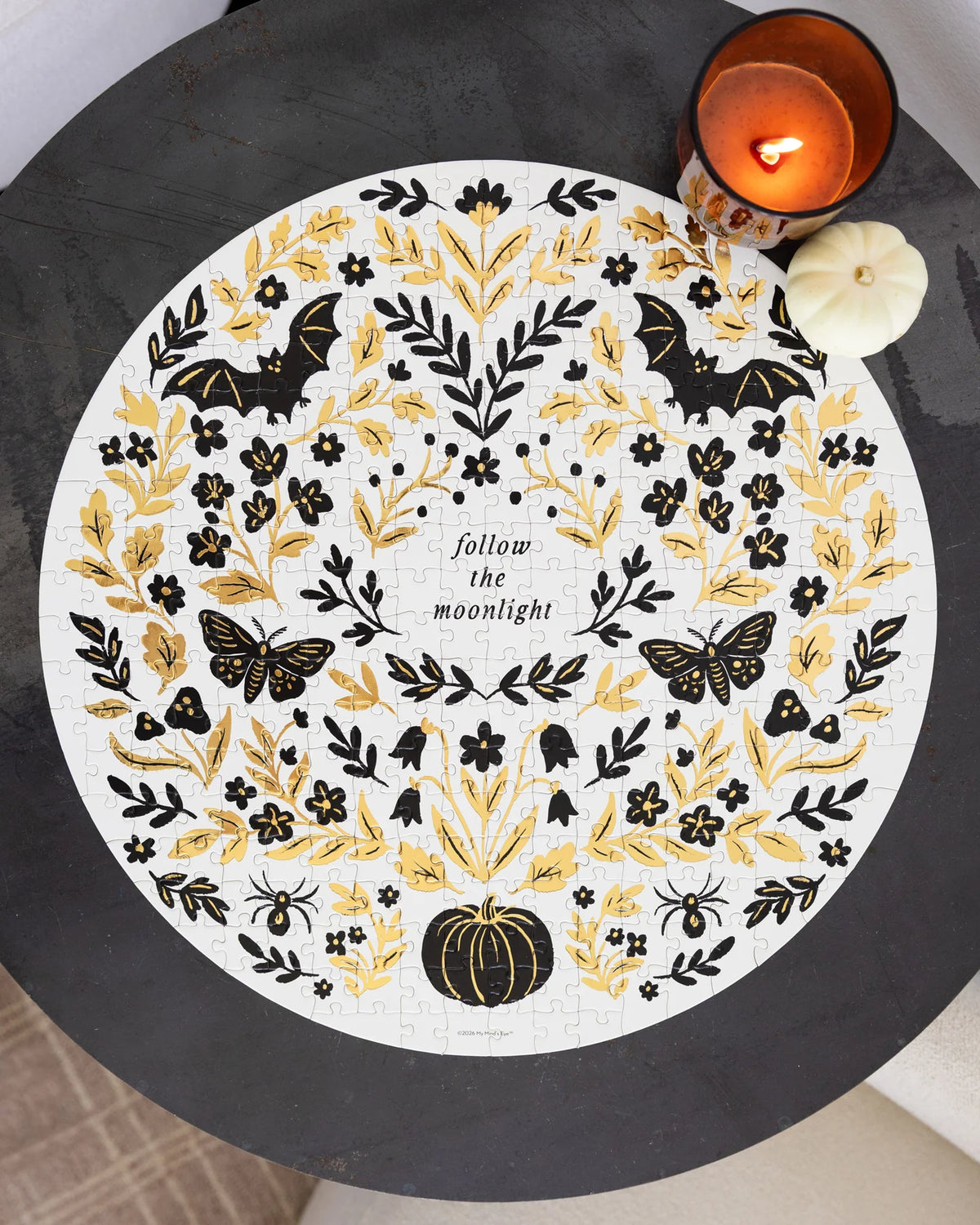 A round white jigsaw puzzle with a black and gold Halloween design, featuring bats, butterflies, leaves, spiders, a pumpkin, and the text 'follow the moonlight', displayed on a dark round table with a lit candle and a small pumpkin prop.