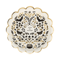 Cream Halloween dessert plate covered in a dense black folk-art pattern of moths, florals, crescent moons, pumpkins, and "follow the moonlight" text, with a gold foil scalloped edge, overhead view.