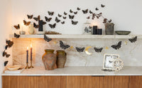 Styled shelf decor featuring the 6 ft moths and stars paper banner strung across a marble shelf alongside black 3D wall moths, terracotta vases, and lit candles.