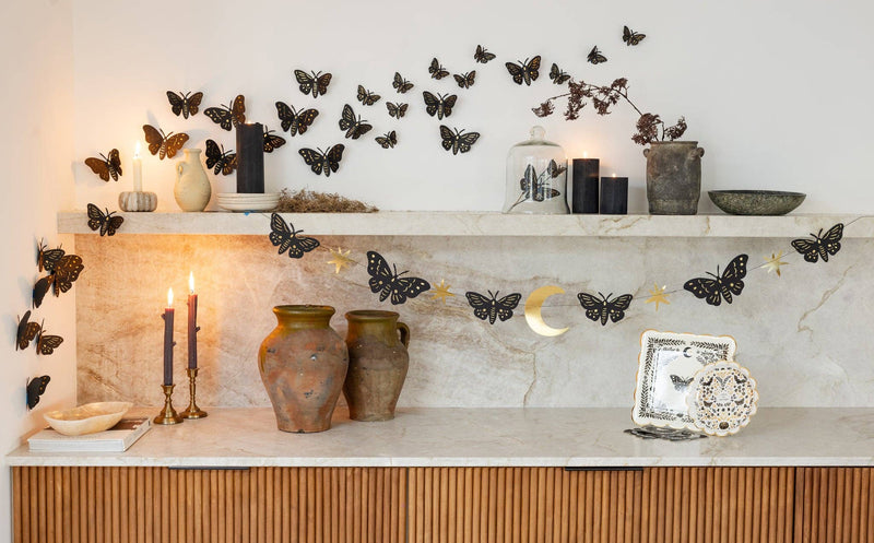 Styled shelf decor featuring the 6 ft moths and stars paper banner strung across a marble shelf alongside black 3D wall moths, terracotta vases, and lit candles.