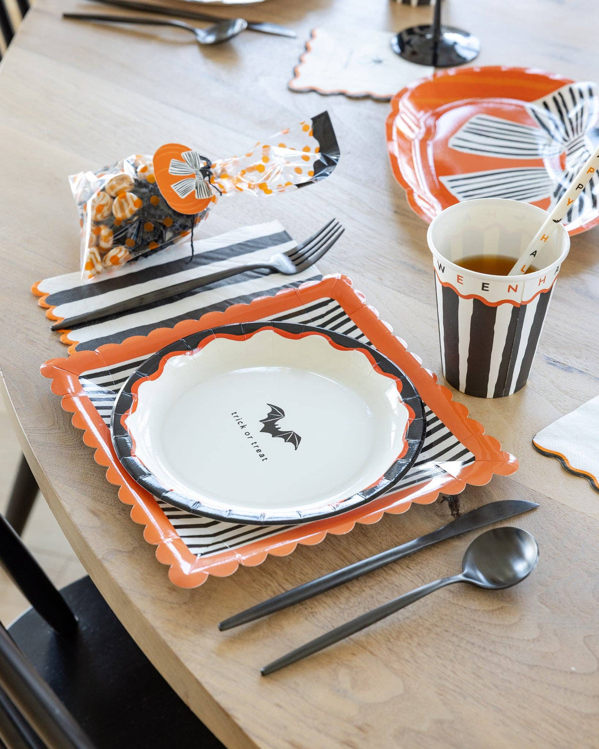 Halloween party place setting showing the spooky icon plate layered on a striped square plate with a striped napkin, HPH cup, cello treat bag, and party hat on a wood table.