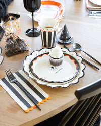 Halloween party place setting showing the plate layered with a smaller spider plate, black-and-cream striped napkin, striped cup, party hat, and treat bag on a wood table.