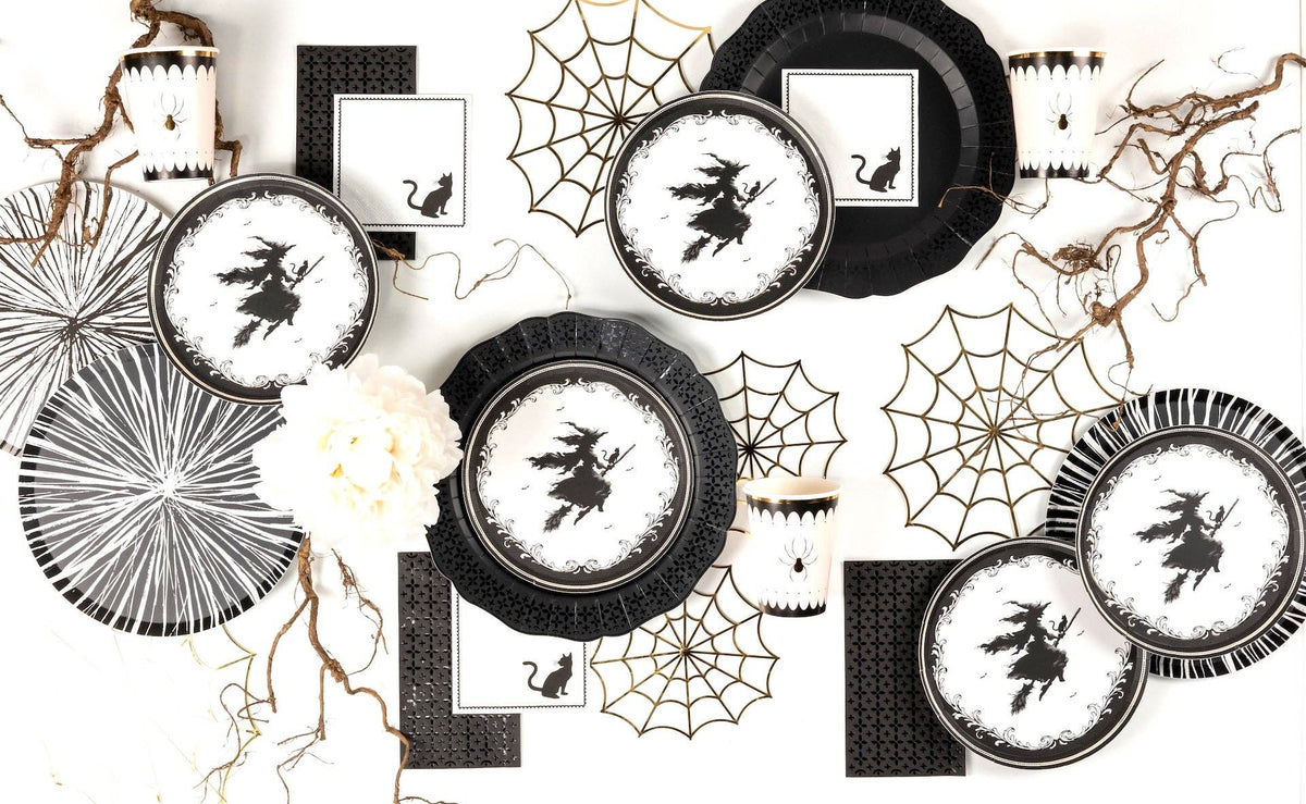 Flat lay of scattered black lace border plates, vintage witch dessert plates, and decorative spider webs on a white surface.