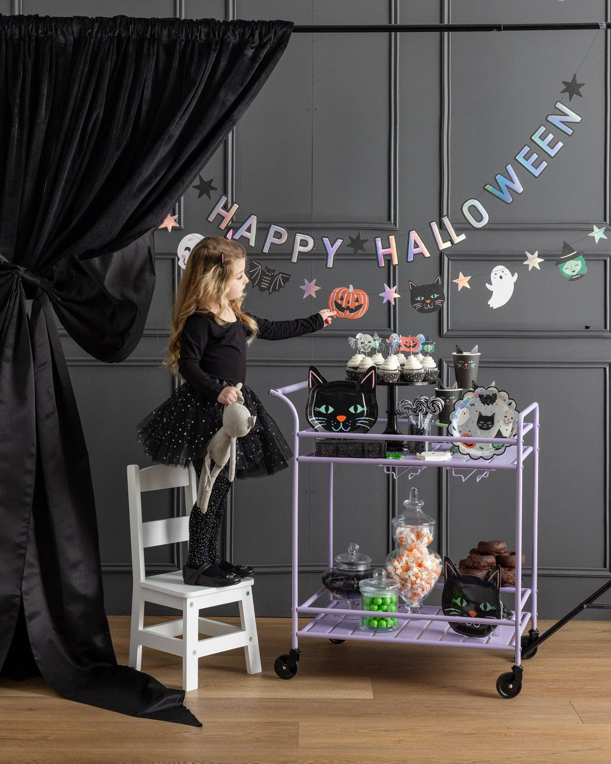 Wide styled shot of the full Halloween party scene — holographic banner on the wall, purple treat cart, black curtains, balloon garland, woman and child posing.