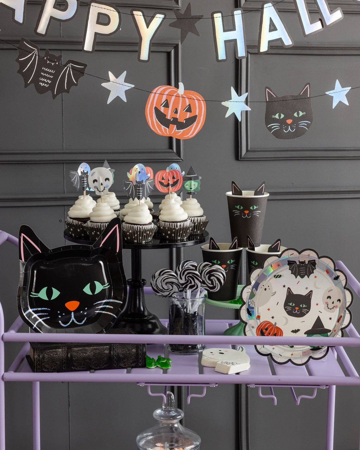 Full Halloween party scene — holographic banner on the wall, purple treat cart styled with cat plates, character cupcakes, ghost and bat hanging decorations, and SPO collection pieces.