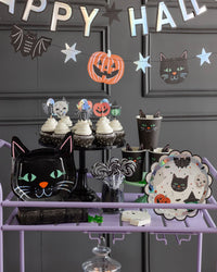 Full Halloween party scene — holographic banner on the wall, purple treat cart styled with cat plates, character cupcakes, ghost and bat hanging decorations, and SPO collection pieces.