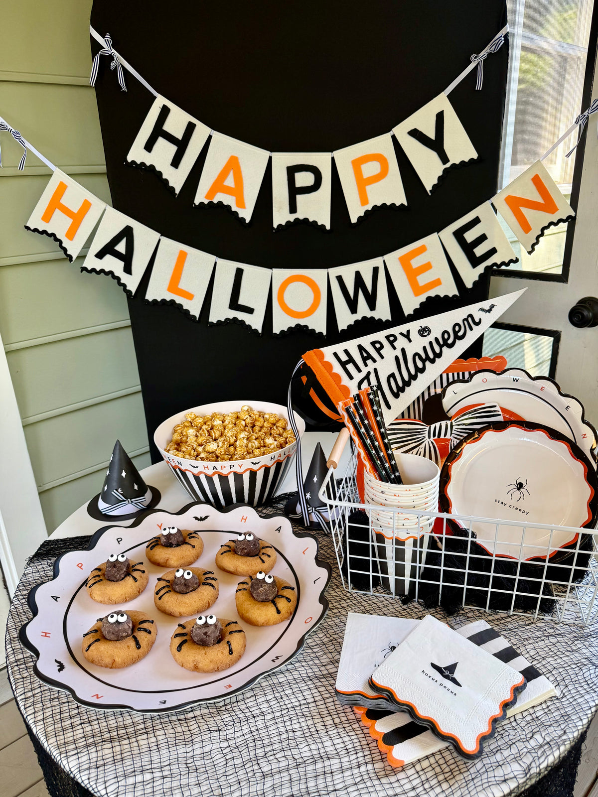 Cream felt Happy Halloween banner with black and orange letters hung above a Halloween party table with spider cookies and caramel popcorn