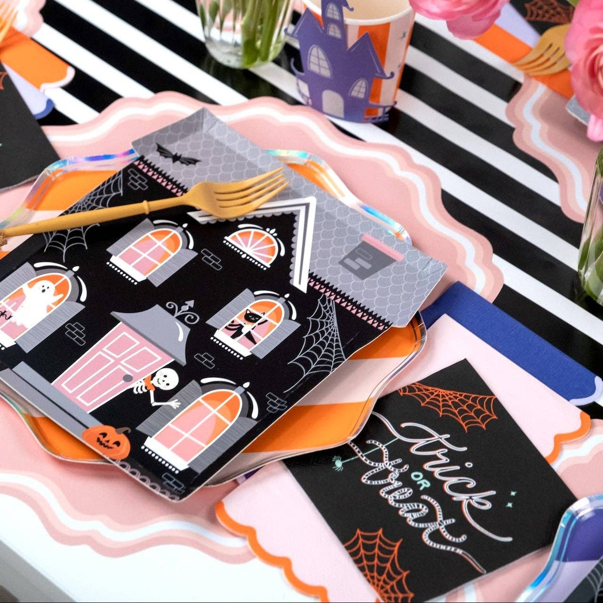 Styled Halloween party table showing the trick or treat napkin with witch-hat plates, orange accents, and pink-and-black elements on a striped surface.