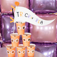 Close-up of “Trick or Treat” halloween pennant perched in a stack of pumpkin cups against shiny balloon tiles, highlighting centerpiece styling ideas.