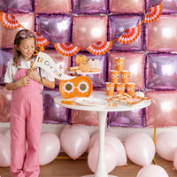 Little girl in pink overalls holds the felt halloween pennant beside a pastel balloon wall and pumpkin cups, showing playful scale for kids’ parties.