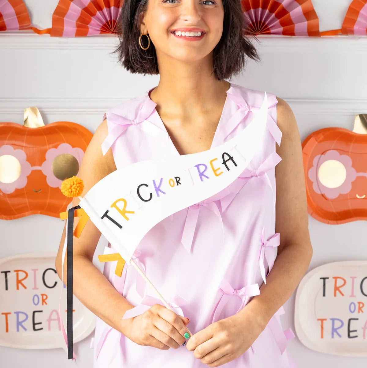 Portrait of smiling woman holding the cream felt halloween pennant with multicolor letters; festive fans and pumpkin plates frame the bright retro vibe.