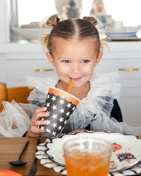 Child holding a star-patterned cup with a star-patterned straw, sitting at a chair.