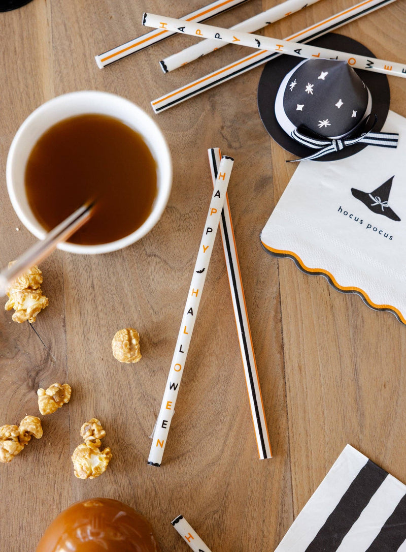 Halloween-themed straws shown close-up (wordmark and stripes on cream) on a wooden table with a cup of coffee, popcorn, and a small witch hat.