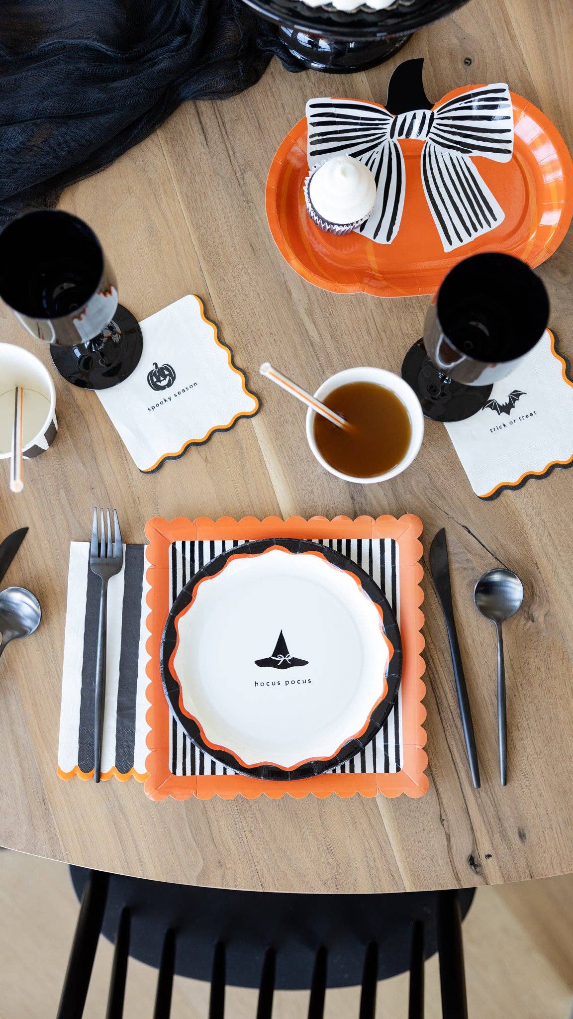 Overhead Halloween table styled shot showing the full HPH layered stack — striped square plate, bow pumpkin plate, and "hocus pocus" icon plate — with cups, cello treat bags, and napkins on a wood table.