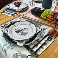 Black and white Halloween toile paper table runner with a cemetery gate, trees and bats under layered toile plates and a fruit board