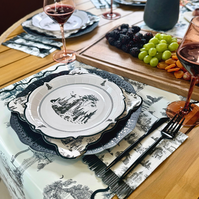 Black and white Halloween toile paper table runner with a cemetery gate, trees and bats under layered toile plates and a fruit board
