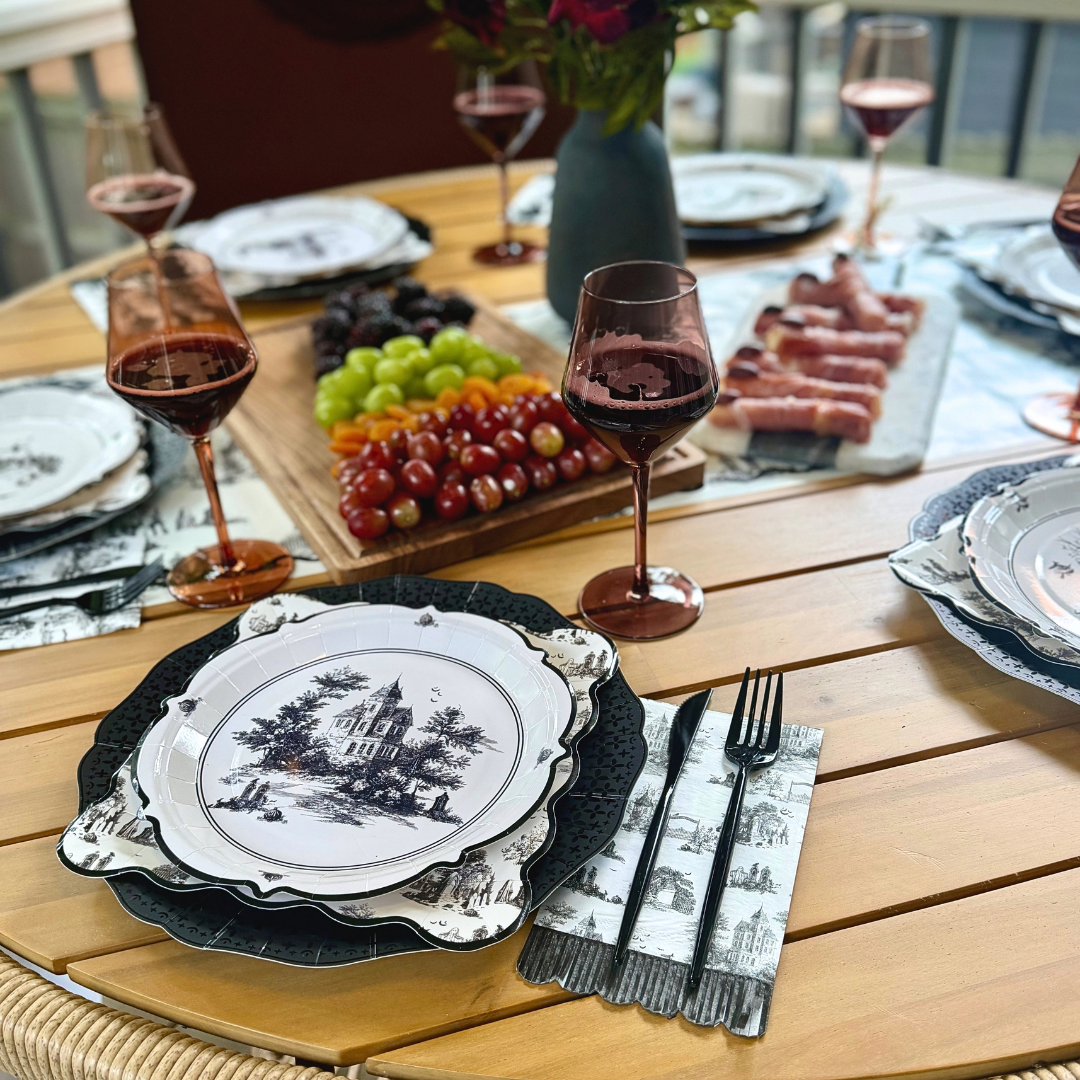 Round Halloween toile paper plate with a haunted house scene on layered plates, with a fringed napkin, black flatware and a charcuterie board behind