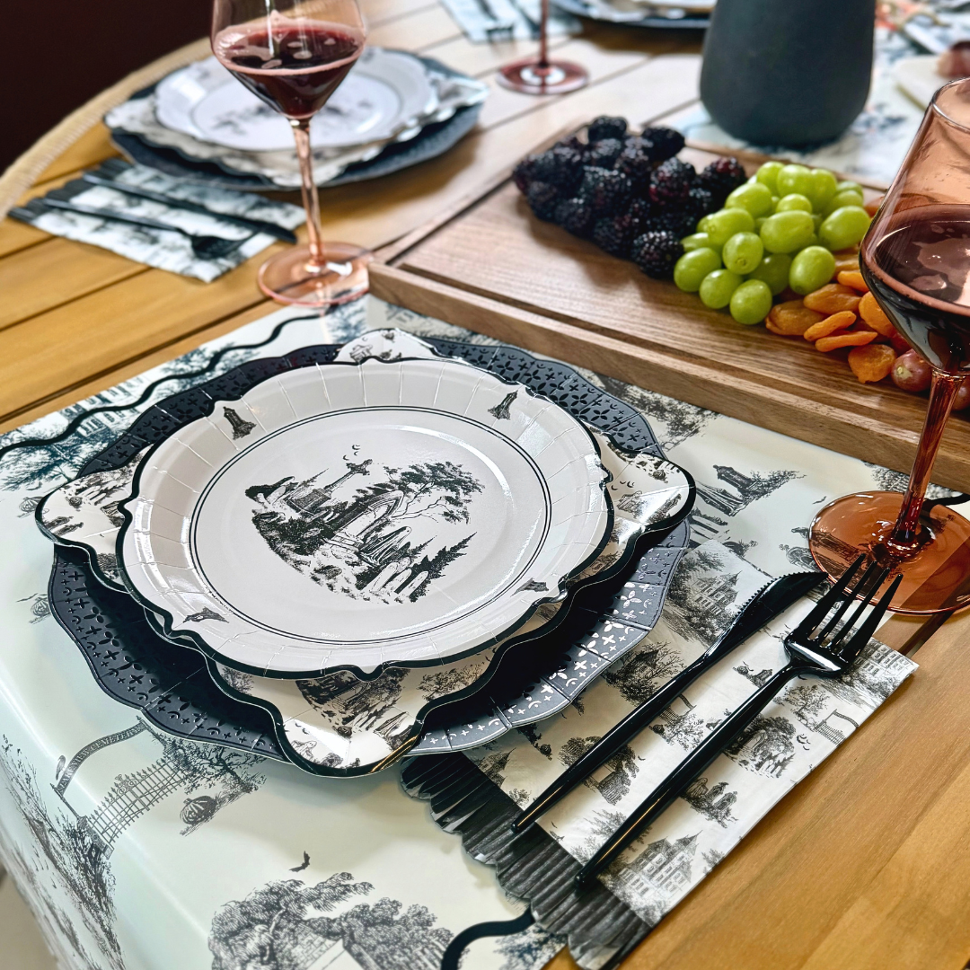Round black and white Halloween toile paper plate with a gothic ruins and graveyard scene, layered on scalloped toile and black plates beside a fruit board