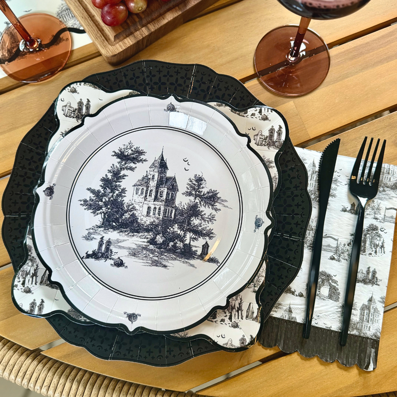 Round Halloween toile paper plate with a haunted house, trees, gravestones and bats in the center and a thin black border