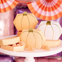 Close crop of orange and white pumpkin shaped treat boxes with gold foil stems surrounded by gumdrops; orange and pink paper fans create a festive backdrop.
