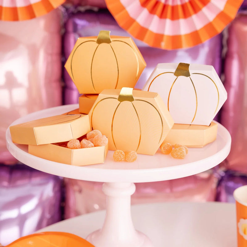 Trio of assembled pumpkin treat boxes—two orange, one white—sit open on a white cake stand spilling gummy candies, framed by retro pink and orange party fans.